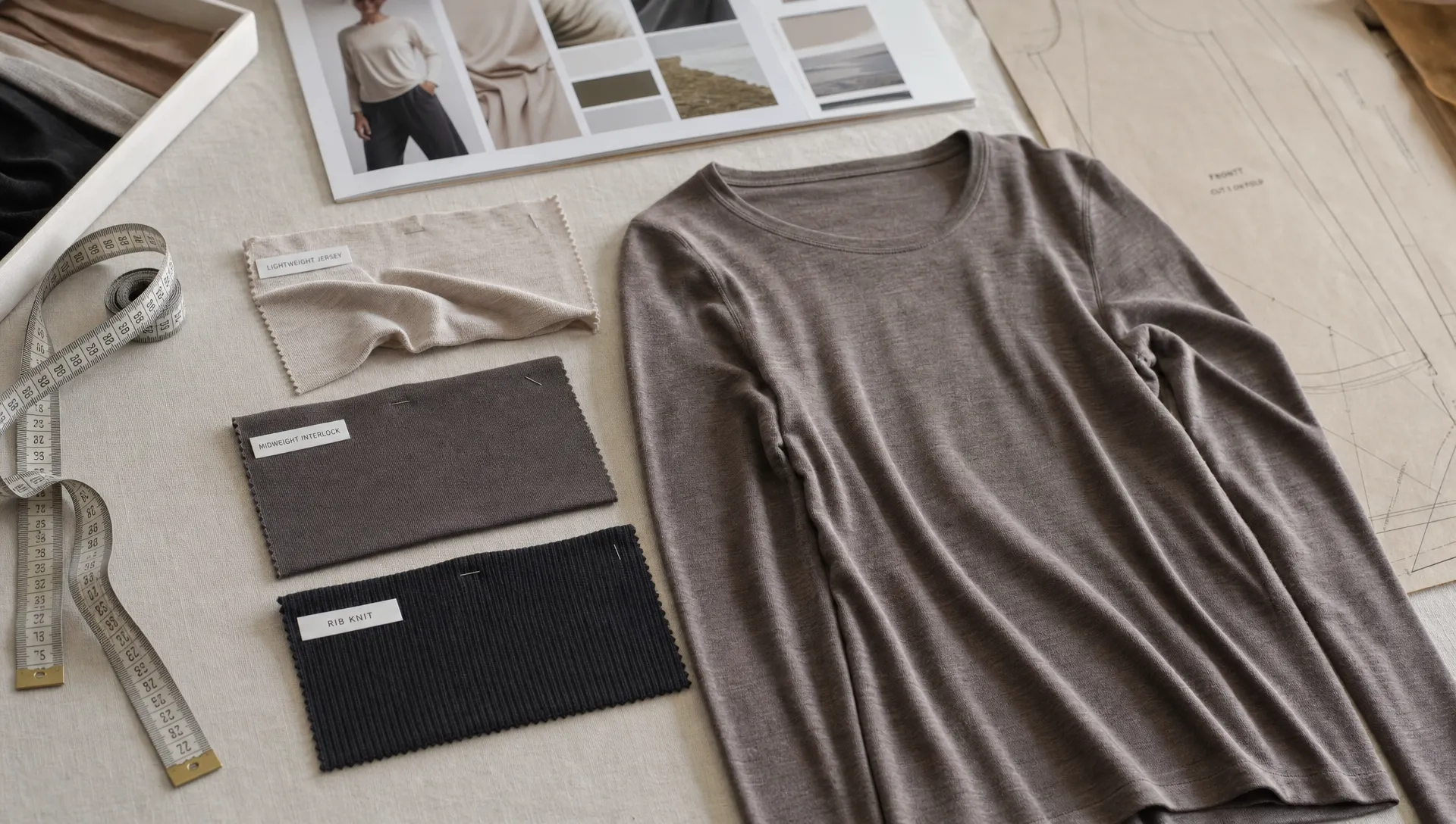 A fabric development table for a merino wool shirt womens collection shows swatches of lightweight jersey, midweight interlock and rib knit beside a measuring tape, pattern pieces and a fit sample.