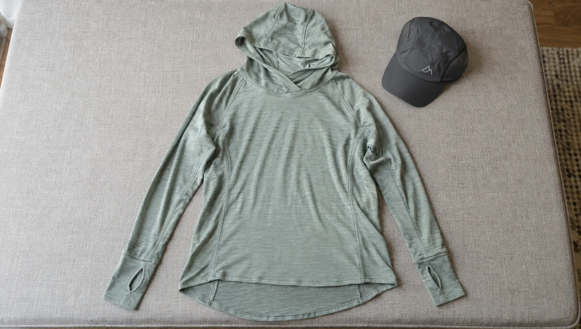 A merino sun hoody lies flat on a fitting platform, showing its shaped hood, overlapping neckline, extended thumbhole cuffs and longer back hem beside a hiking cap.