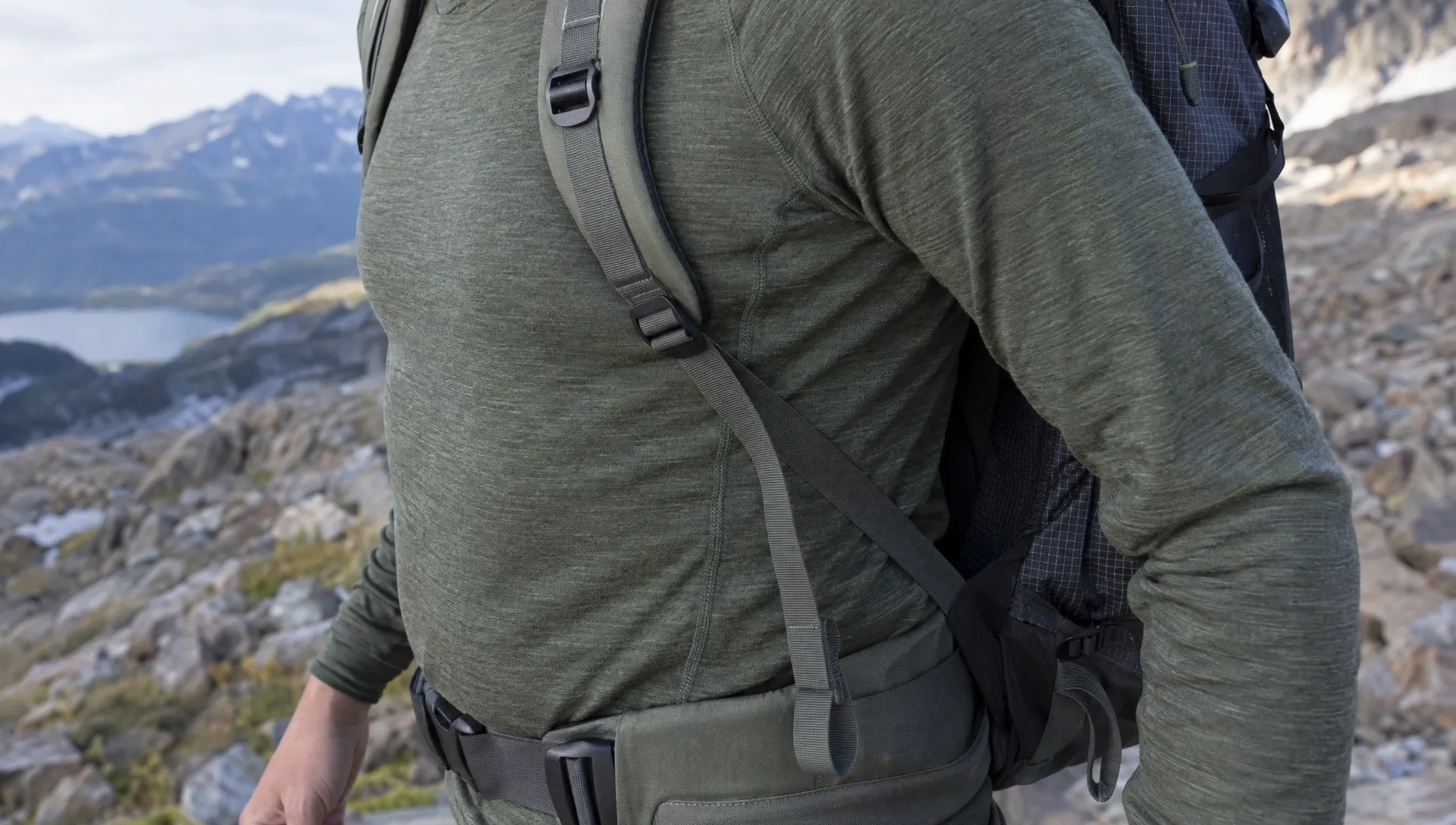 A merino wool base layer worn beneath a backpack, with the waist belt contacting the fabric and the underarm seam and cuff visible for wear inspection.