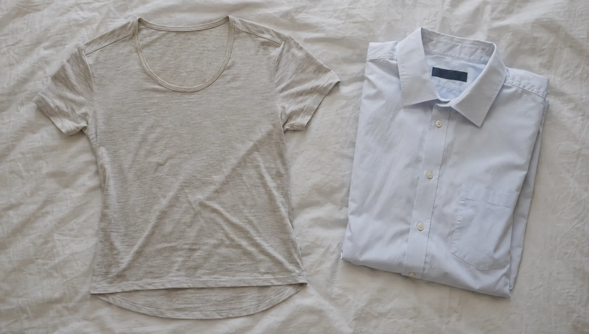 A merino wool undershirt lies beside a pale dress shirt, showing its low neckline binding, smooth shoulder seam, and longer back hem.