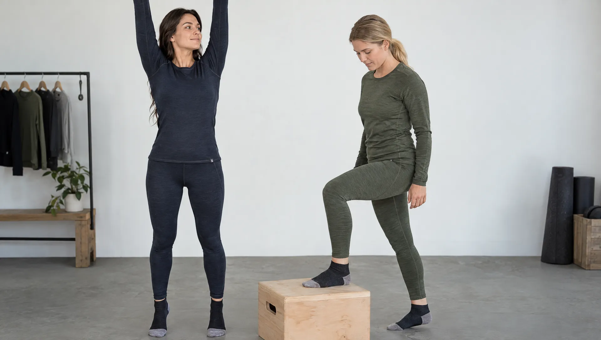 Two adult women test merino base-layer fit in a studio: one raises both arms while the other steps onto a low platform, showing how the tops and leggings move.